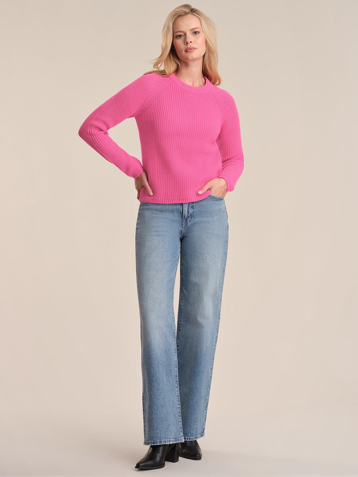 A person stands against a plain background wearing a 525 America bright pink cotton shaker stitch sweater with raglan sleeves, light blue straight-leg jeans, and black heeled boots, one hand in a pocket and the other on their hip.