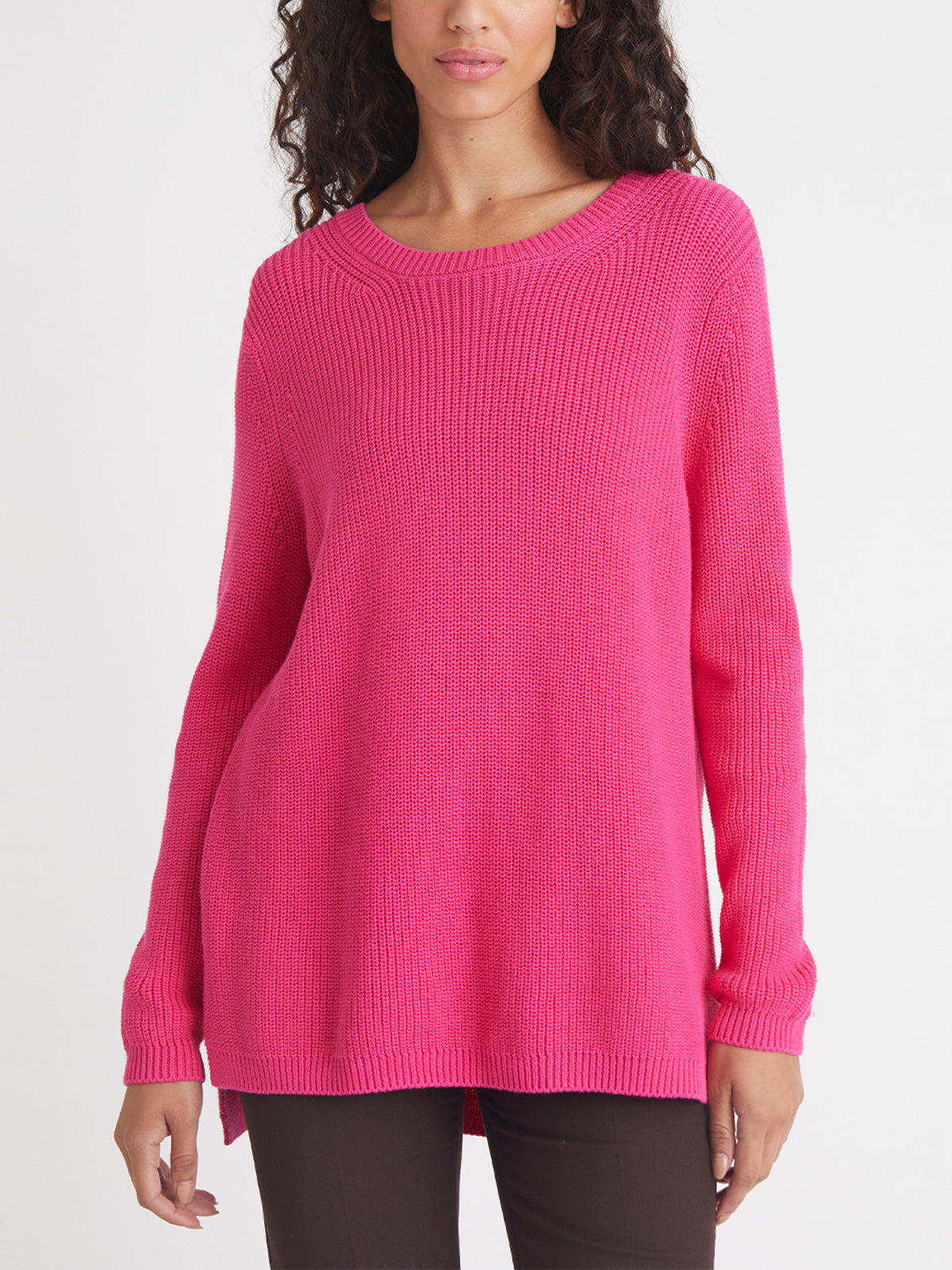 A woman stands against a white background wearing a 525 America relaxed fit bright pink 100% cotton knit sweater with long sleeves and brown pants.