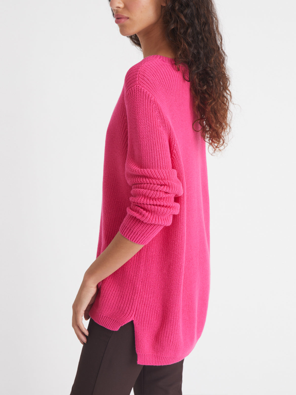 A woman stands in profile wearing a relaxed fit, bright pink 100% cotton knit sweater with long sleeves by 525 America and black pants against a plain white background.