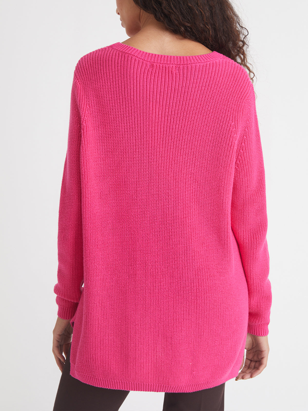 A person with curly hair is shown from the back wearing a relaxed fit, long-sleeved, bright pink shaker stitch sweater by 525 America and dark pants against a plain white background.