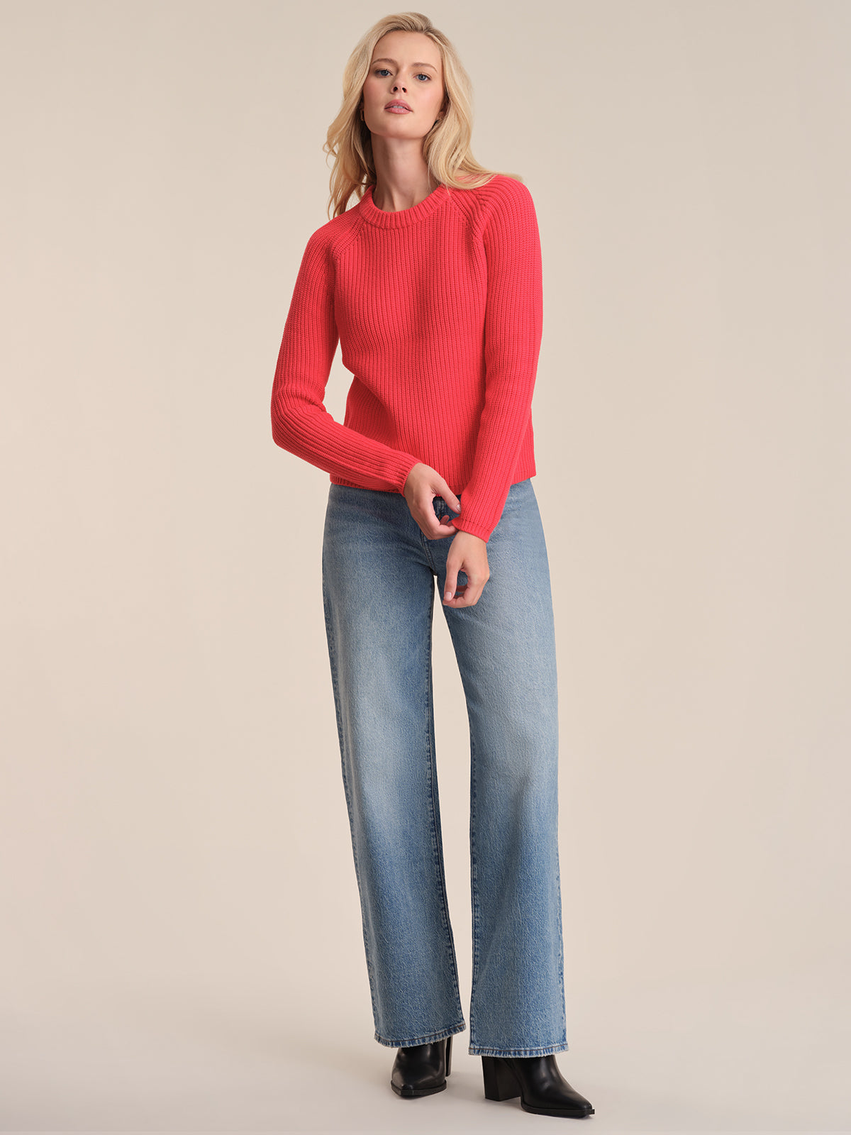 A person with blonde hair stands against a light background, wearing a bright red 525 America cotton shaker stitch sweater with raglan sleeves, light blue wide-leg jeans, and black boots, looking neutrally at the camera.