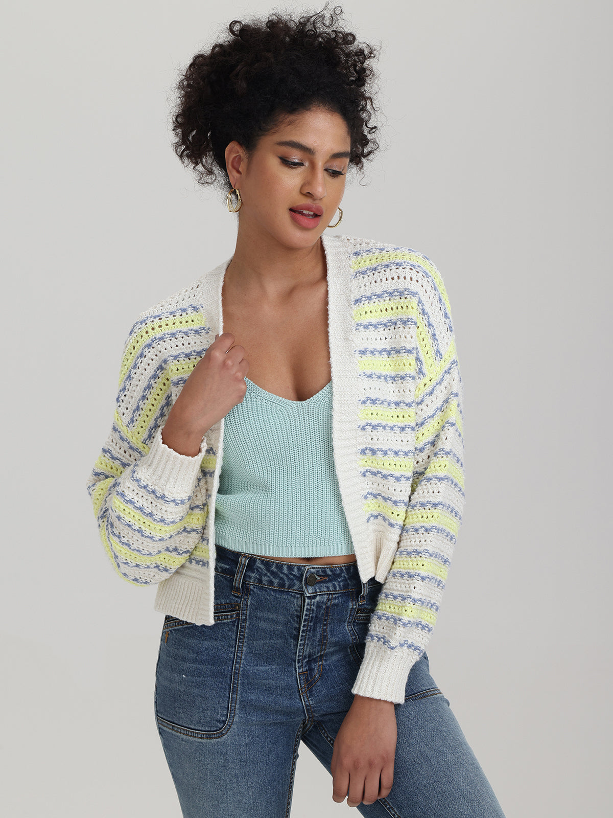A person with curly hair is modeling a striped cardigan by 525 America over a light blue top and relaxed-fit blue jeans, slightly tilting their head while holding the cardigan open. The background is plain and neutral.