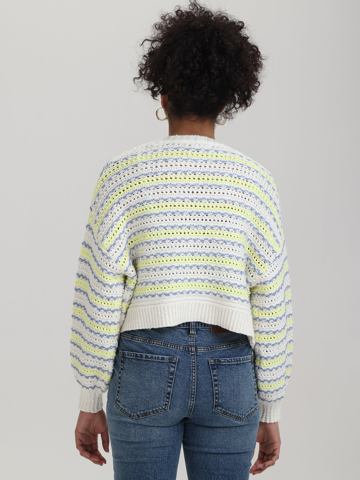 A person with curly hair faces away, wearing a relaxed-fit crocheted sweater by 525 America, with pastel yellow, blue, and white stripes. Theyre in high-waisted blue jeans and hoop earrings against a plain gray backdrop.