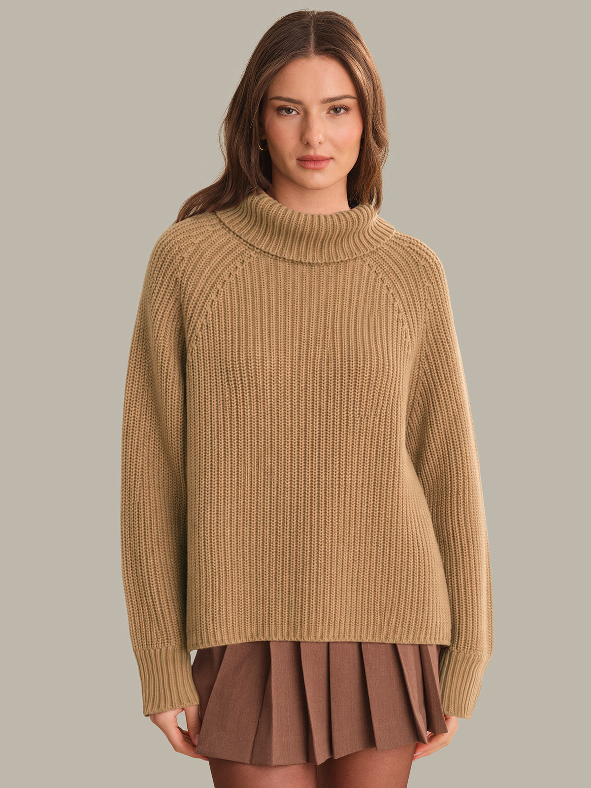 Woman wearing a beige sweater and brown pleated skirt on a plain background