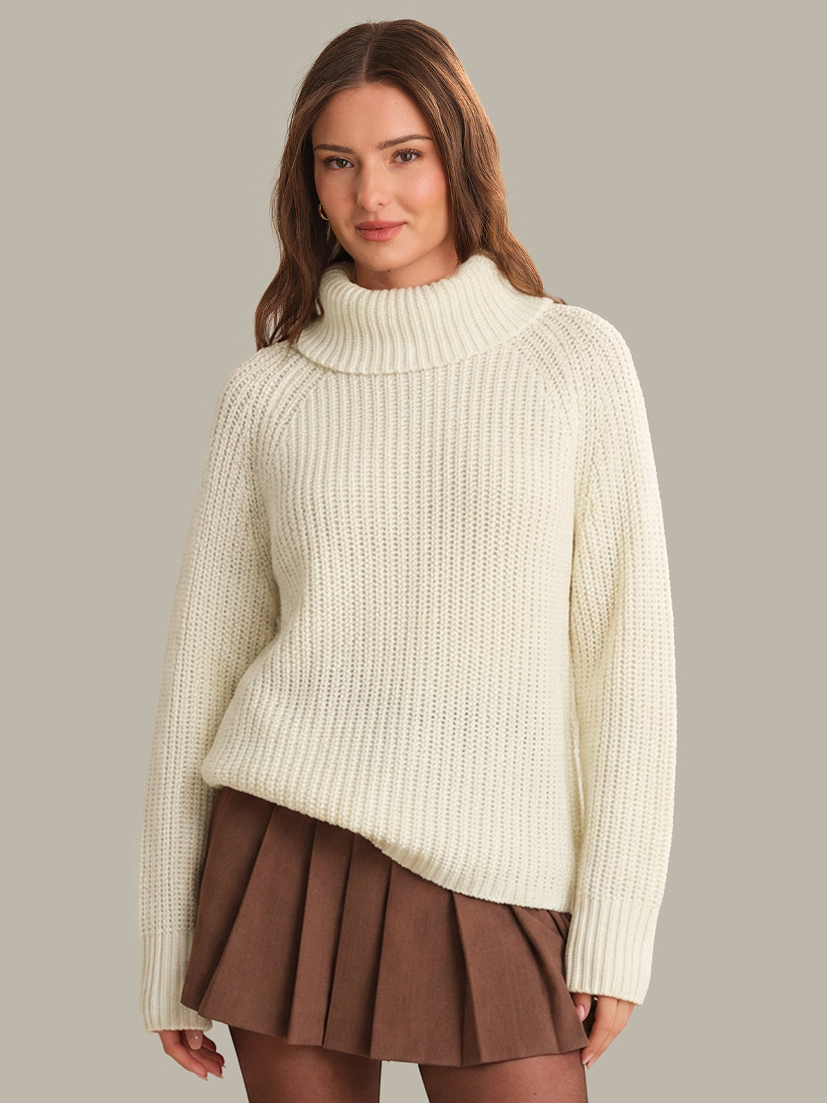 Woman wearing a cream knitted sweater and brown pleated skirt on a plain background