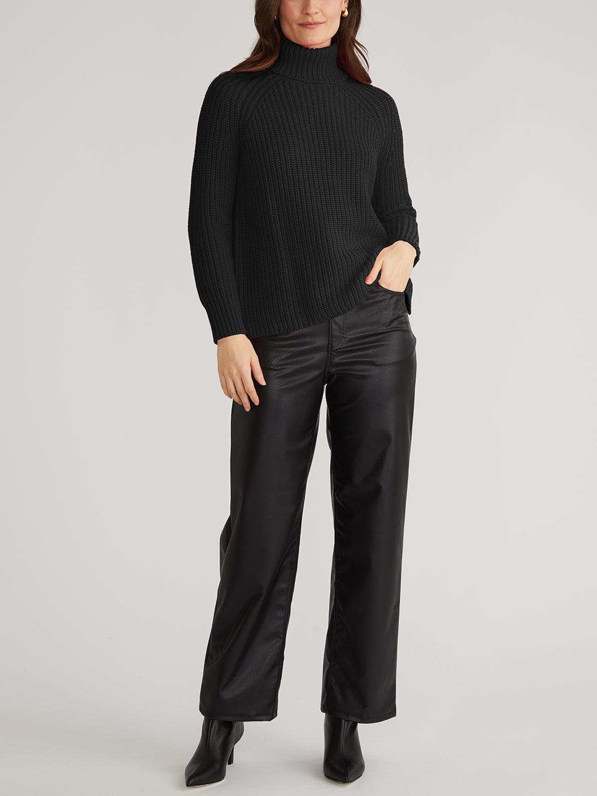 A person in a cozy black ribbed turtleneck sweater by 525 America and black leather pants stands against a plain background. They have one hand in their pocket, paired with black ankle boots.