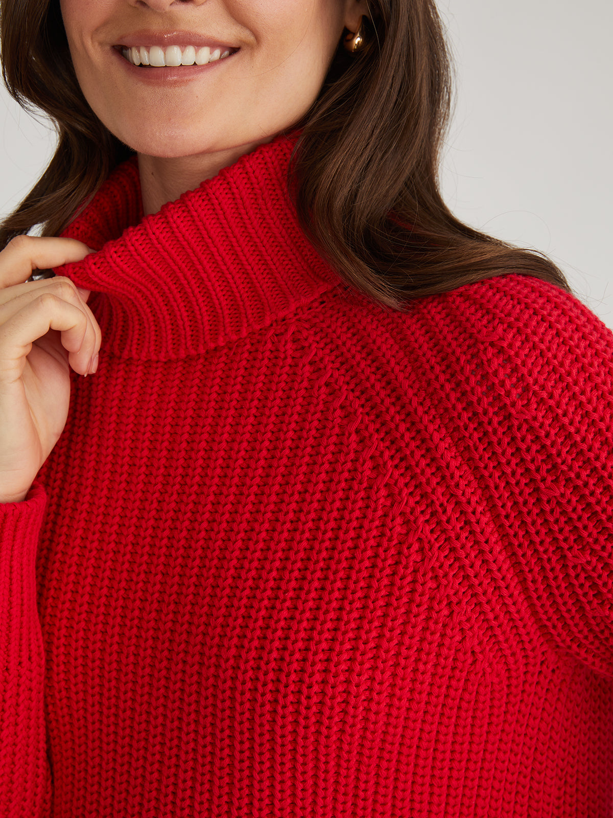 A person wearing a vibrant red 525 America turtleneck sweater smiles warmly. The 100% cotton fabric features a textured knit pattern, with their long brown hair partially visible against a plain, neutral background, enhancing the cozy vibe.