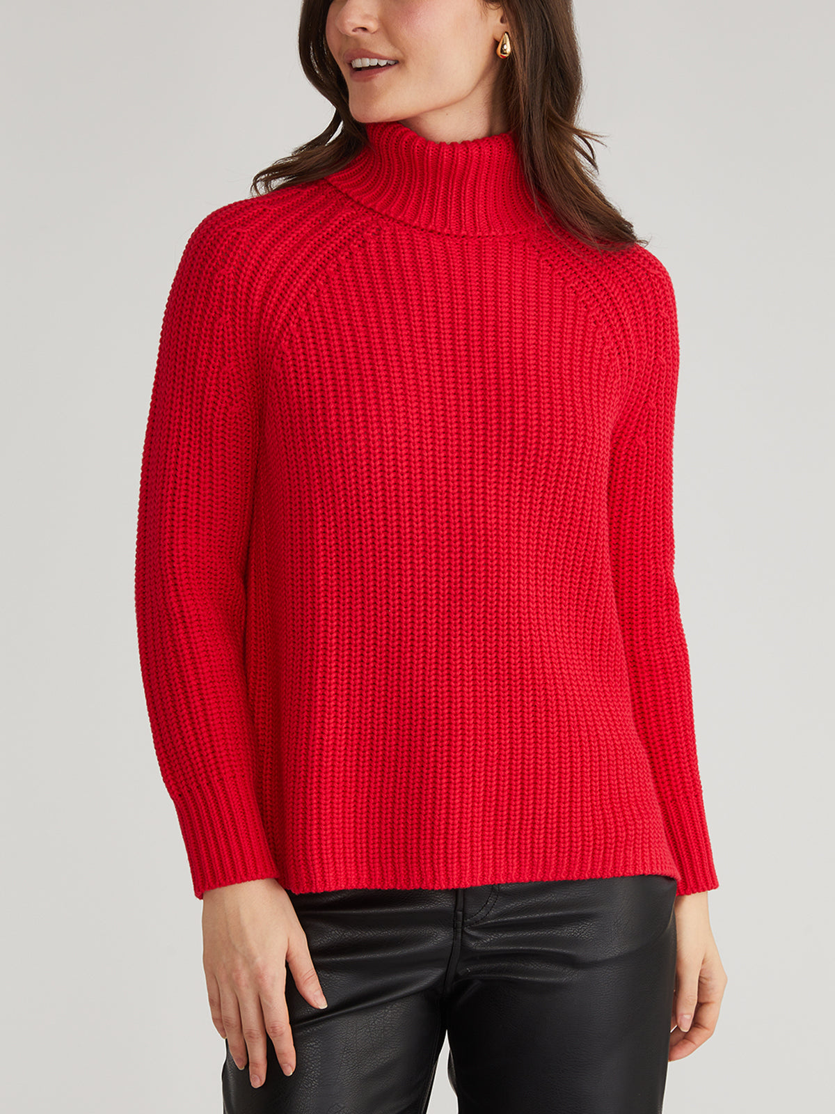 A person wearing a vibrant red 525 America turtleneck sweater stands with their left hand on their thigh. They have long brown hair and wear black leather-style pants against a plain light gray background.