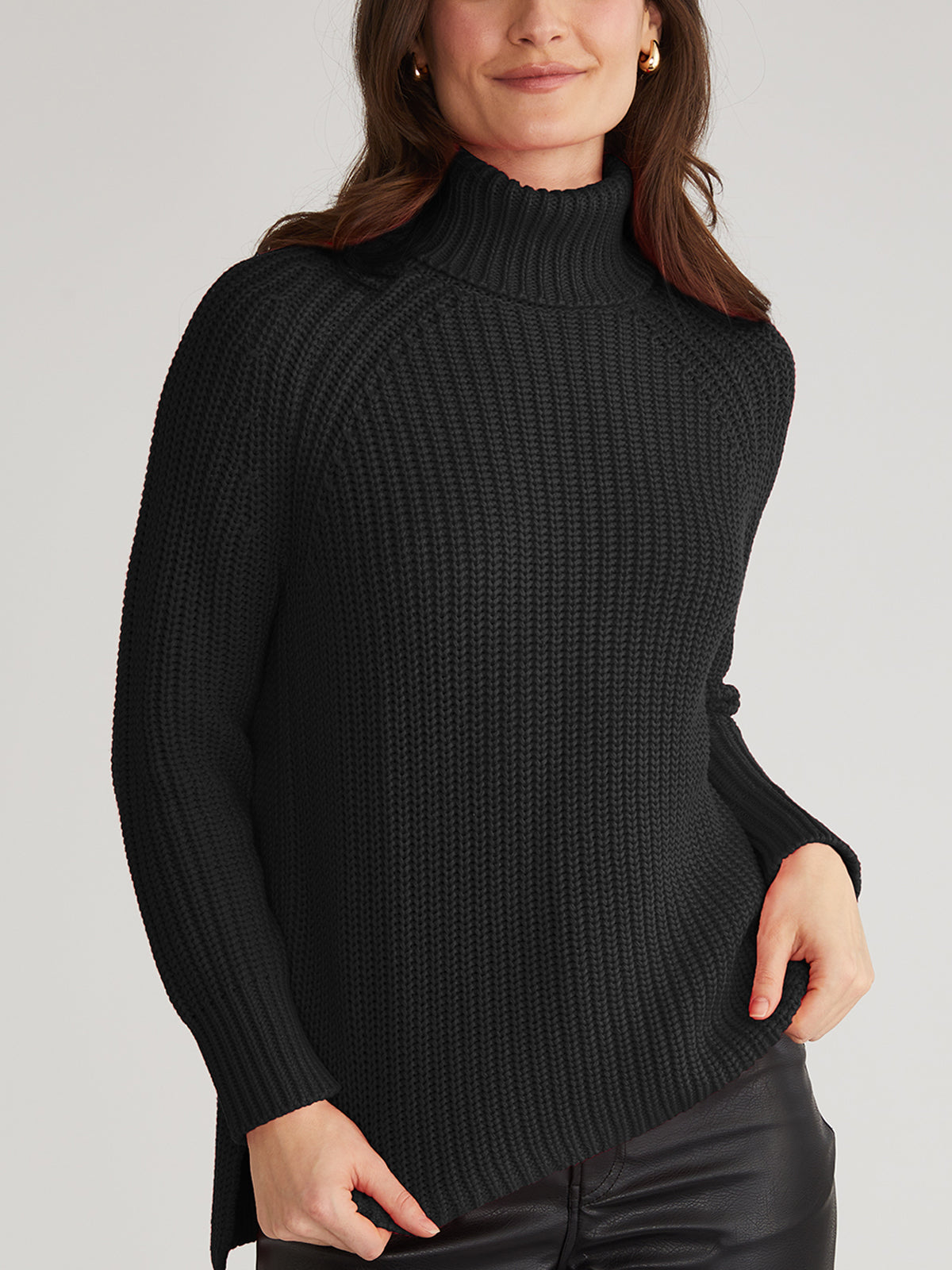 A person in a comfy, black ribbed turtleneck sweater from 525 America, crafted from 100% cotton, paired with black pants stands against a plain background with their right hand tucked in a pocket.