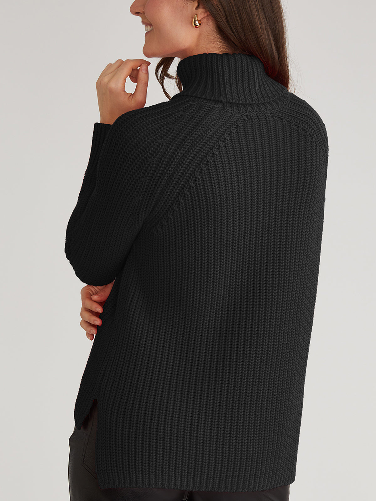 A woman smiles, touching her face with her right hand, while wearing a cozy 525 America black ribbed turtleneck sweater. Made of 100% cotton, it has a high collar and long sleeves. Shes viewed from the back and side against a plain light-colored background.
