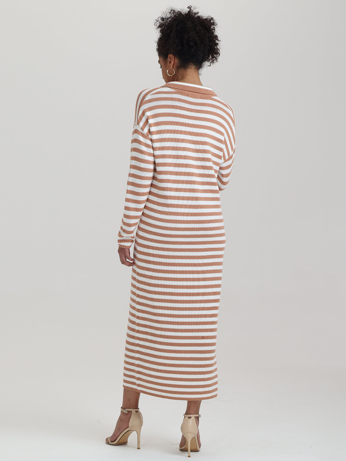 A person with curly hair is wearing a relaxed-fit, ankle-length sailor-stripe dress in tan and white from 525 America. Theyre also sporting beige high-heeled sandals and hoop earrings, standing against a plain white backdrop, ideal for transitional weather.