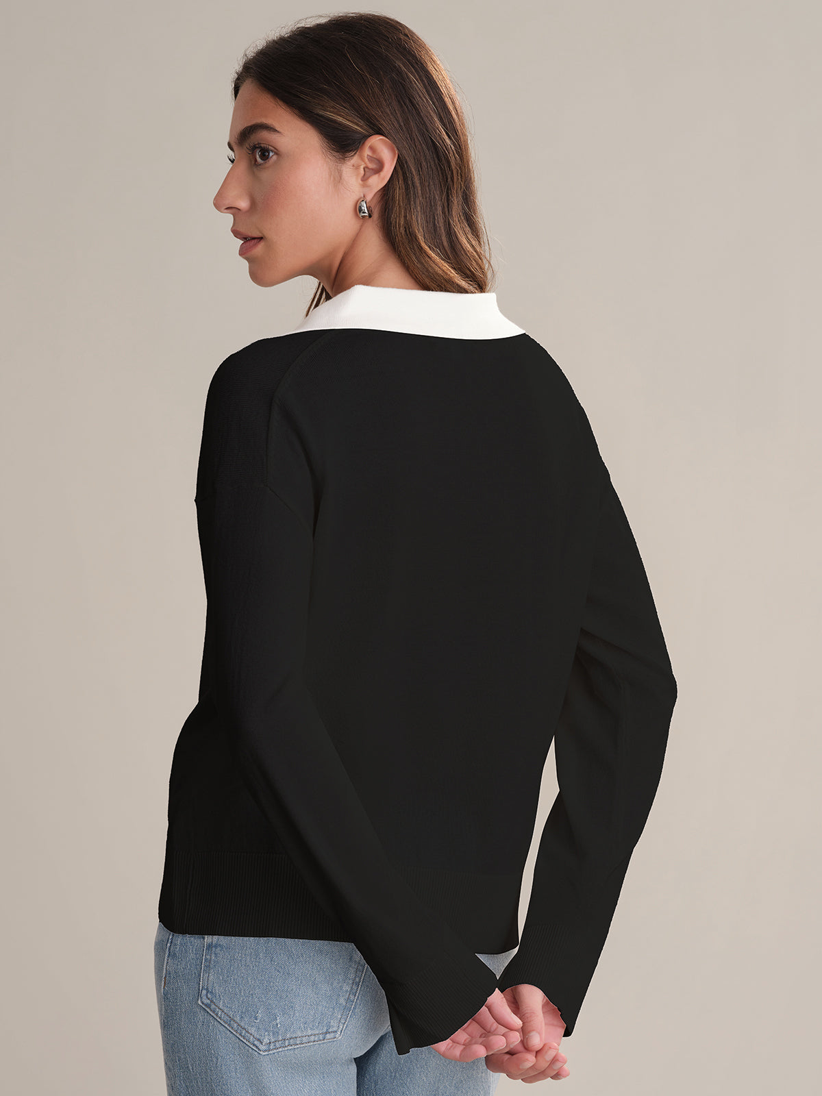 A woman with shoulder-length brown hair, hands clasped behind her back, wears a black 525 America lightweight pullover with a white Johnny collar and light blue jeans. She stands facing away, turning her head to the side.