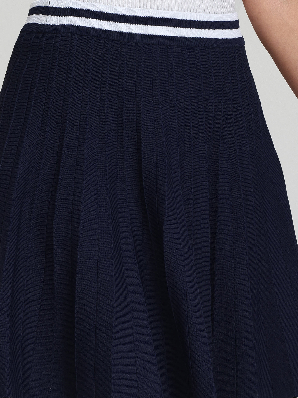 A sporty-chic vibe is captured in this close-up of a person wearing a navy blue pleated skirt by 525 America, featuring intricate pleat details and a striped waist. The textured fabric is displayed prominently.
