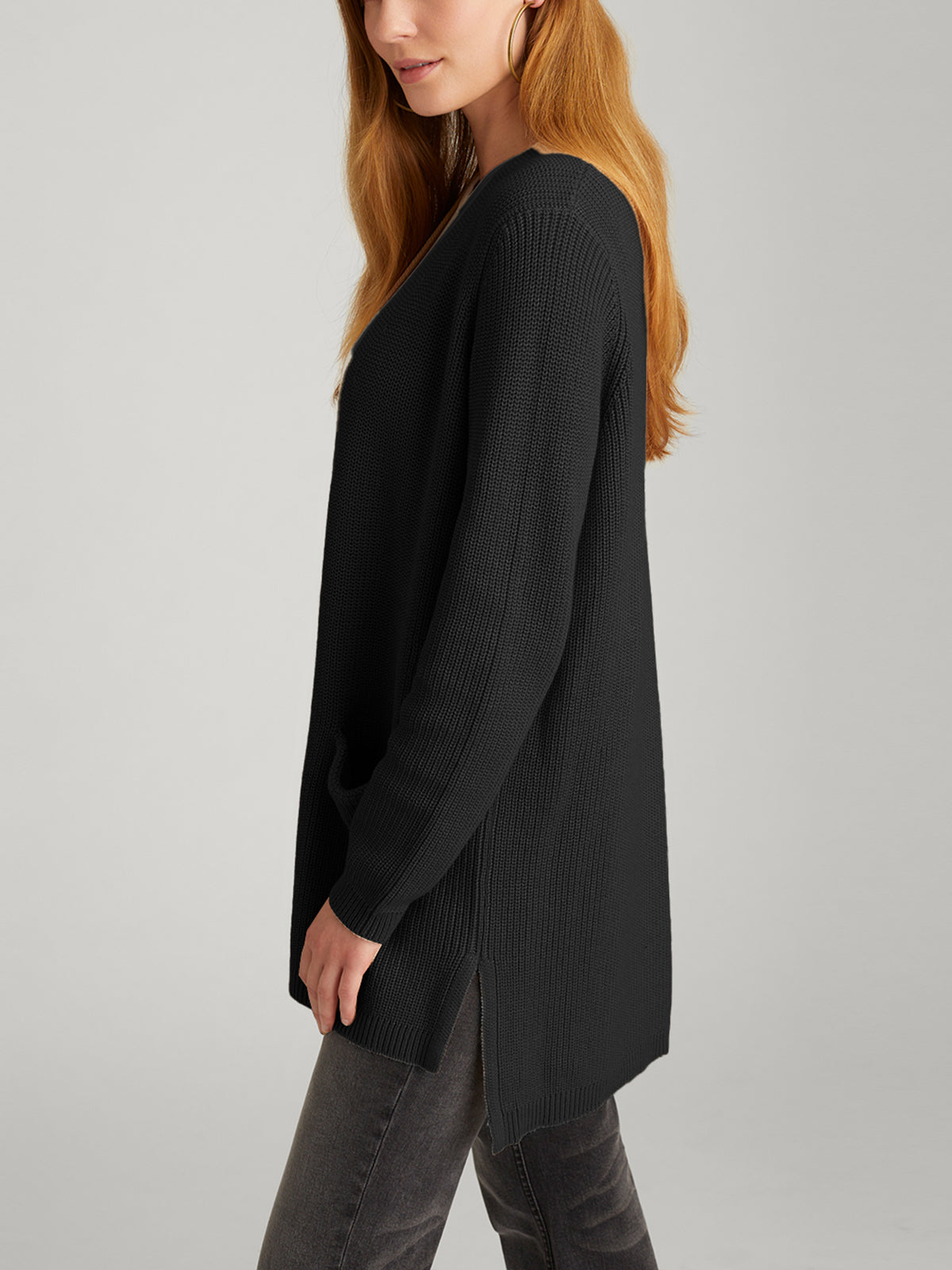 A woman with long hair wears a black, ribbed sweater and dark jeans. She stands sideways with a neutral expression, effortlessly completing her look by layering the Emma: Shaker Stitch Open Cardigan over the ensemble against a plain gray background.