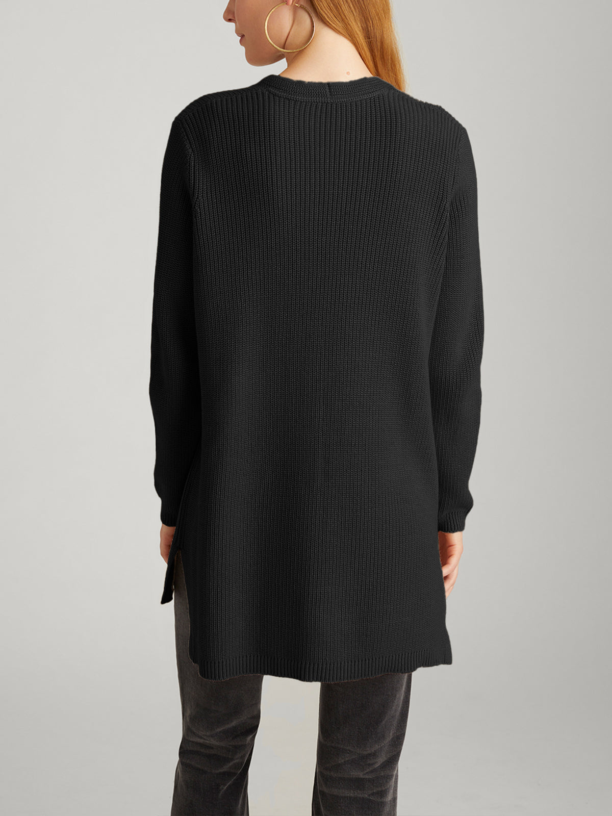 Someone with long hair is wearing the stylish black Emma: Shaker Stitch Open Cardigan along with dark pants. They sport large hoop earrings and are set against a neutral gray background, facing away.