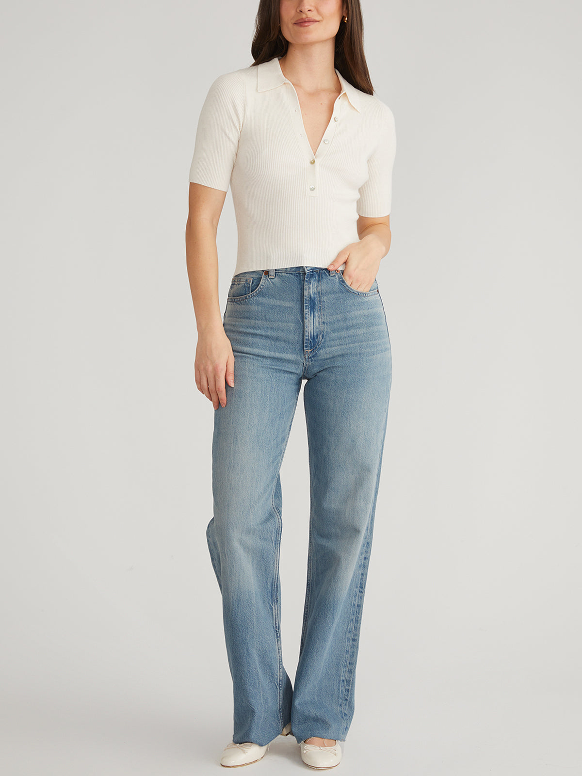 A person poses against a plain backdrop in a chic outfit from 525 America: a short-sleeve white ribbed top with a Johnny collar, light blue wide-leg jeans, and stylish white shoes, with one hand casually in their pocket.
