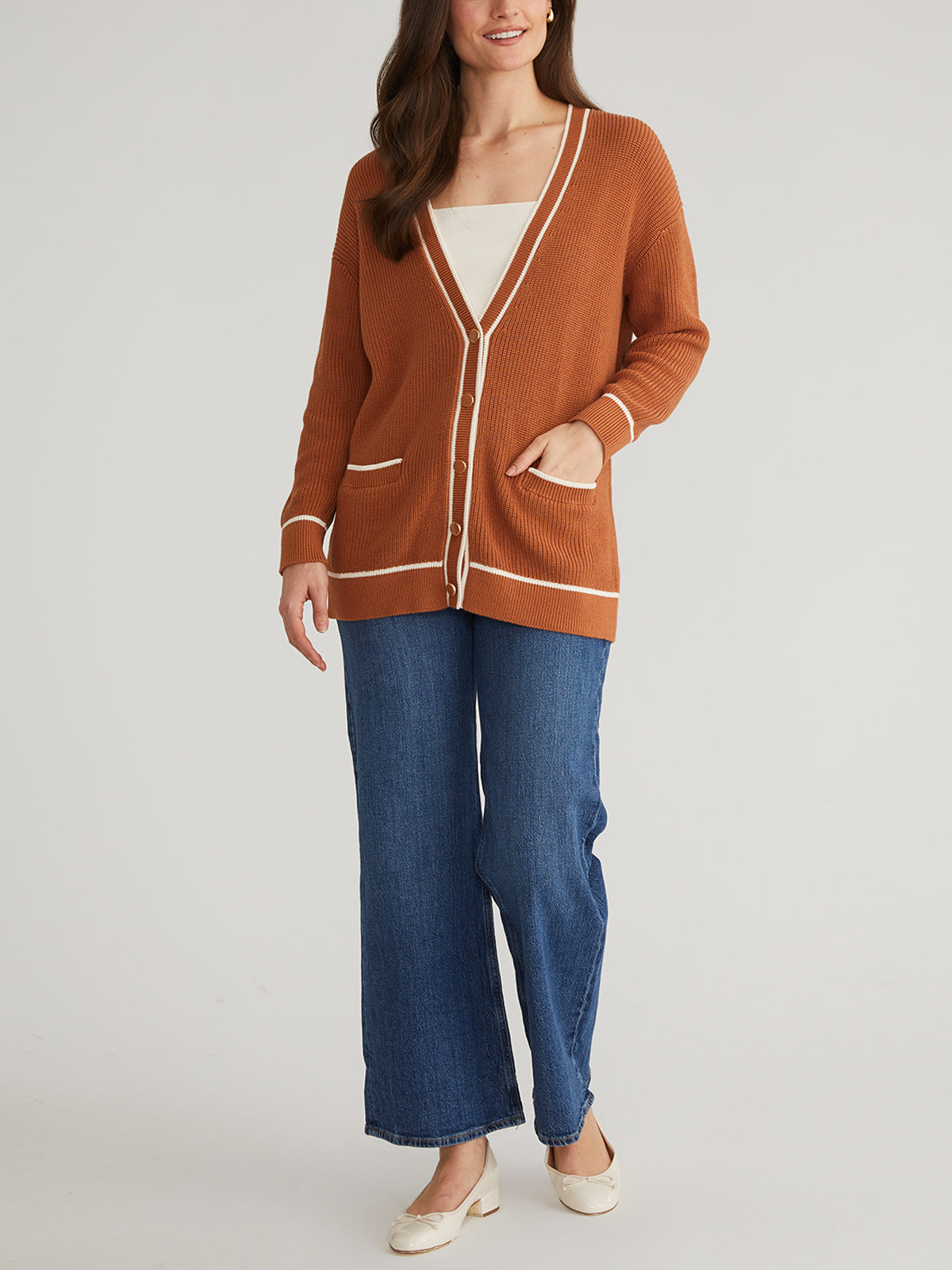 A woman with long brown hair stands against a plain backdrop wearing a 525 America relaxed fit, grandpa-style cardigan with white trim over a white top, paired with blue wide-leg jeans and white shoes, hands tucked in her cardigan pockets.