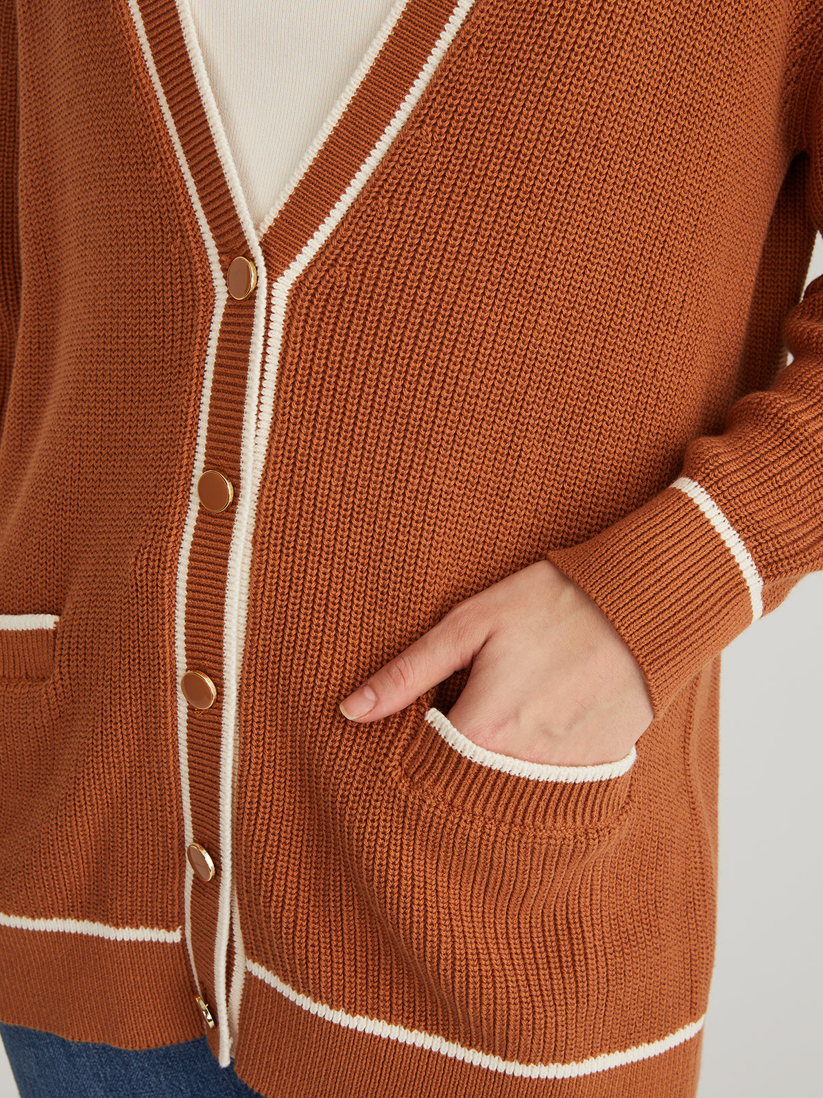 Wearing a 525 America relaxed-fit cardigan in rust with white trim and gold buttons, they have one hand in the front pocket. Paired with blue jeans, a light-colored shirt completes the look.