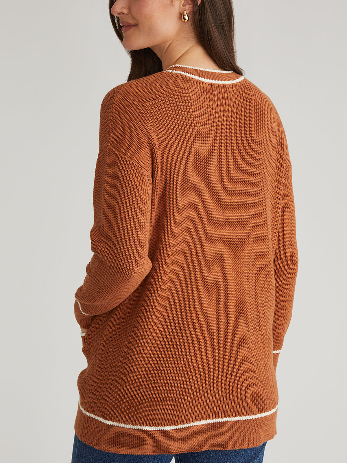 A person wears a brown grandpa-style cardigan with white trim from 525 America. The relaxed fit complements their long dark hair as they pose with one hand in their pocket against a plain, light-colored background.