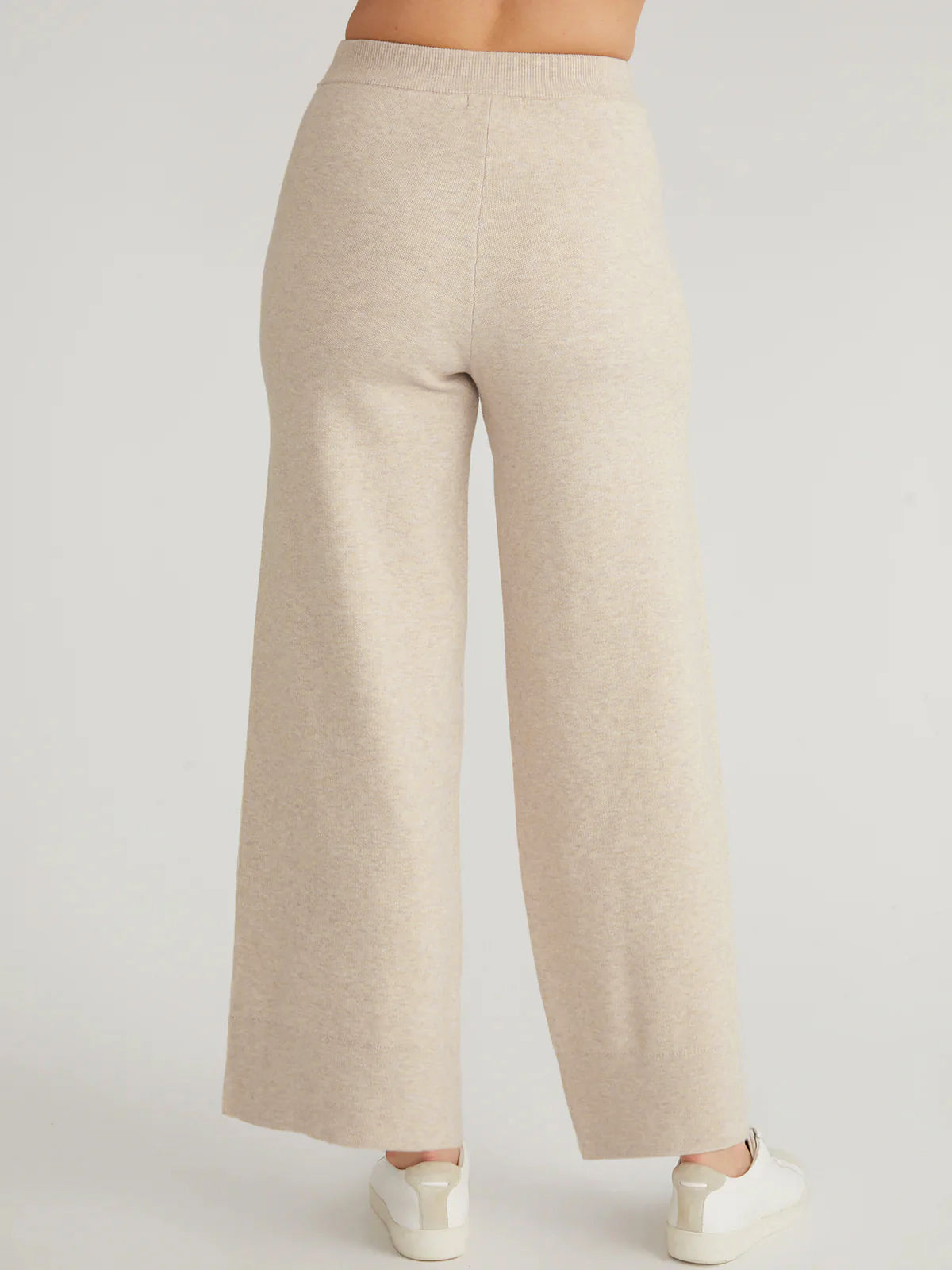 The back view features a person in 525 Americas loose, beige high-rise wide-leg pants paired with white sneakers. The torso is obscured, highlighting the relaxed silhouette as they elegantly stand on a light-colored surface.
