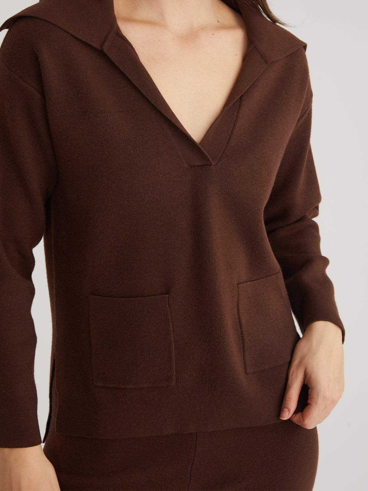 Someone is styled in a chic 525 America piece—a brown V-neck long-sleeved top with two front pockets. Their right hand rests on their hip against a light, plain background, making it ideal for enhancing any winter wardrobe.