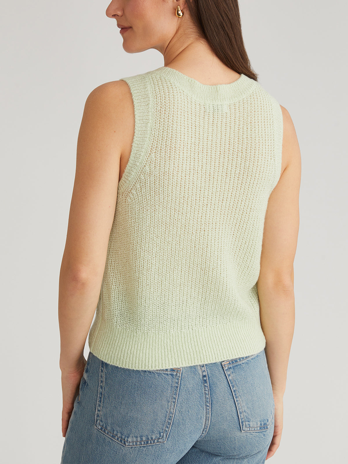 Wearing a sleeveless light green knit top from 525 America, featuring pointelle stitching, and blue jeans, a person stands with their back to the camera against a plain light-colored background.