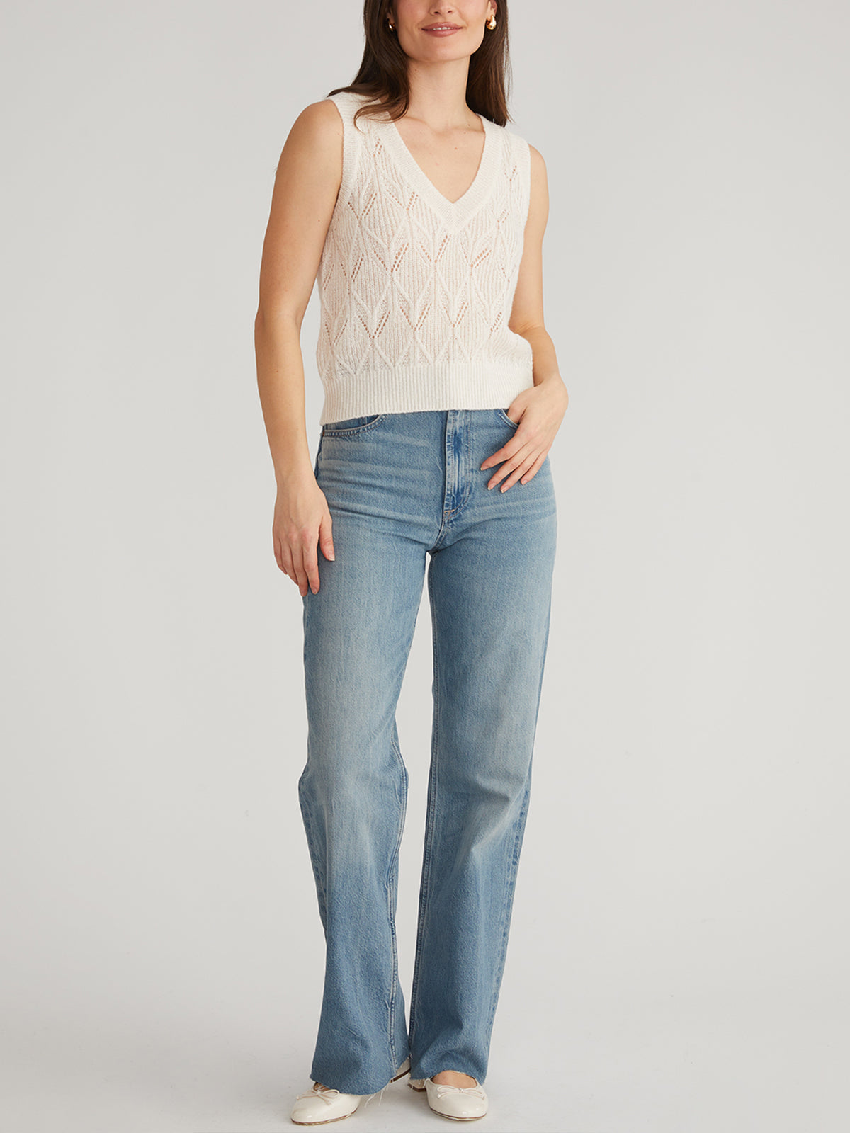 A person poses against a plain background, wearing a cream sleeveless knit top with pointelle stitching by 525 America, light blue jeans, and light-colored sneakers. One hand rests in their pocket.