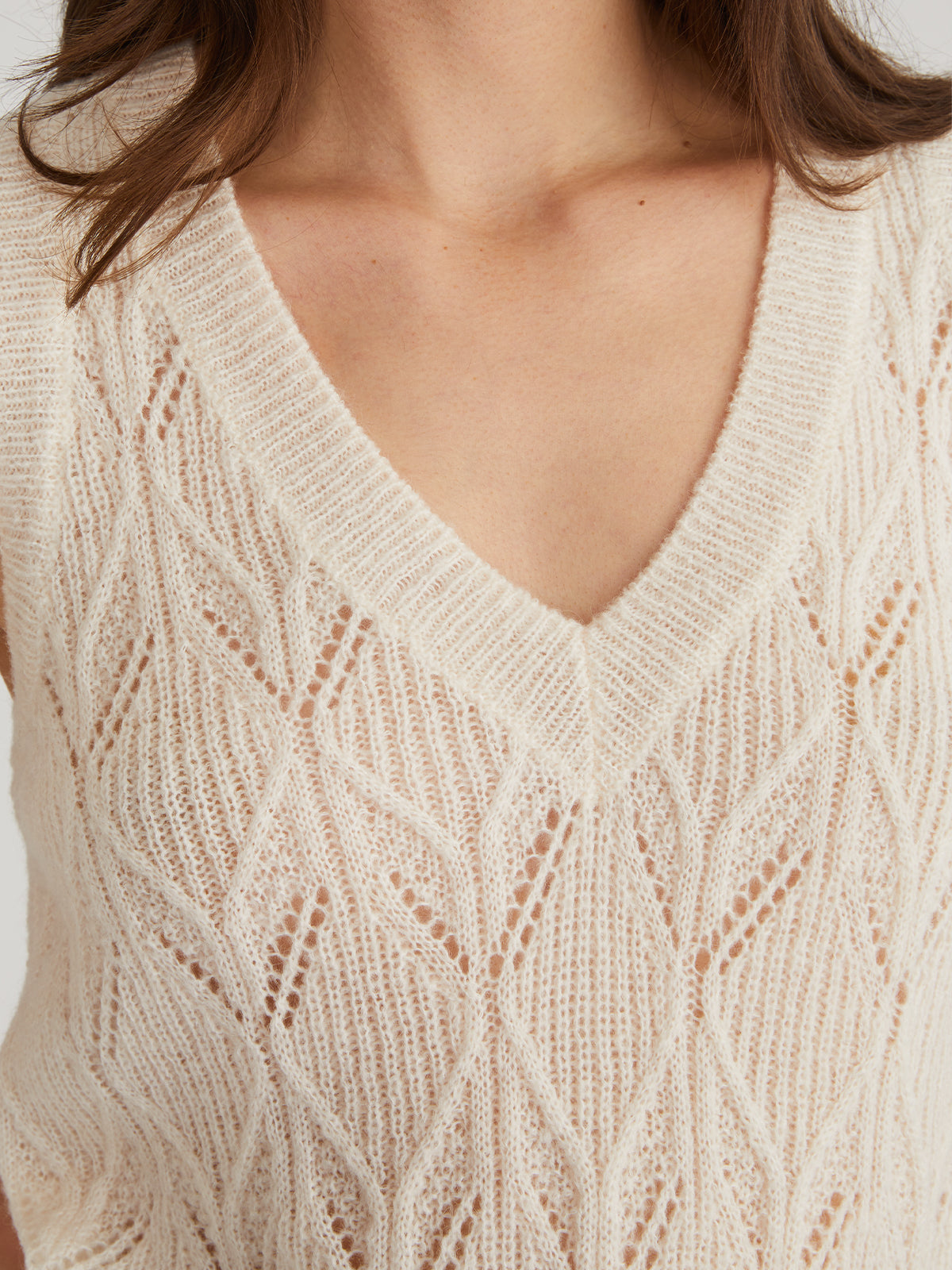 A chic look features a person in a sheerer, cream V-neck sweater vest by 525 America, boasting pointelle stitching and a geometric pattern, worn against bare skin. Soft hair drapes over the shoulders.