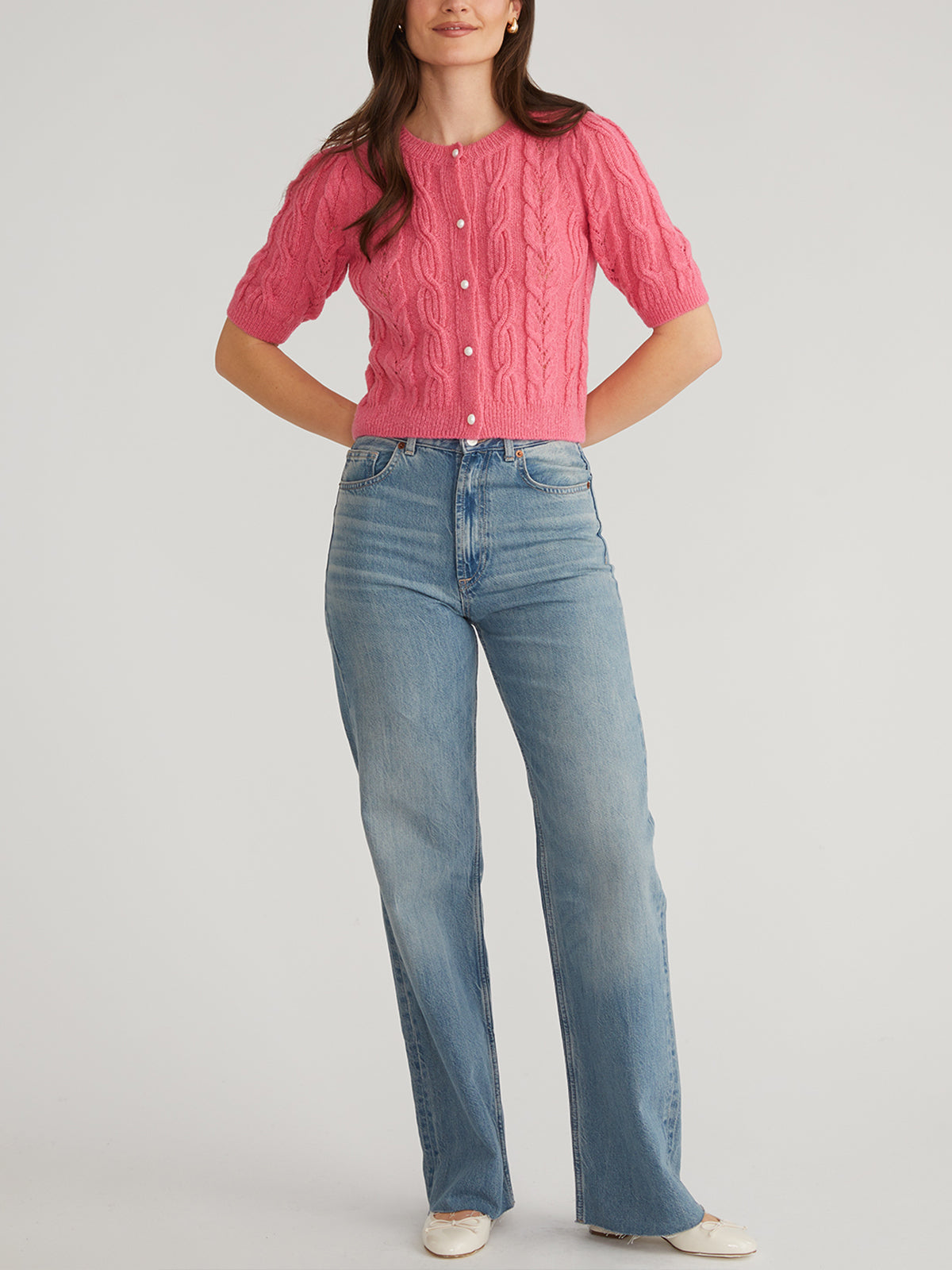 A person wears a pink cable-knit puff short-sleeve sweater from 525 America with pearl-like white buttons and blue high-waisted jeans, posing against a plain light backdrop. Their arms are relaxed at their sides, and white shoes peek out at the bottom.