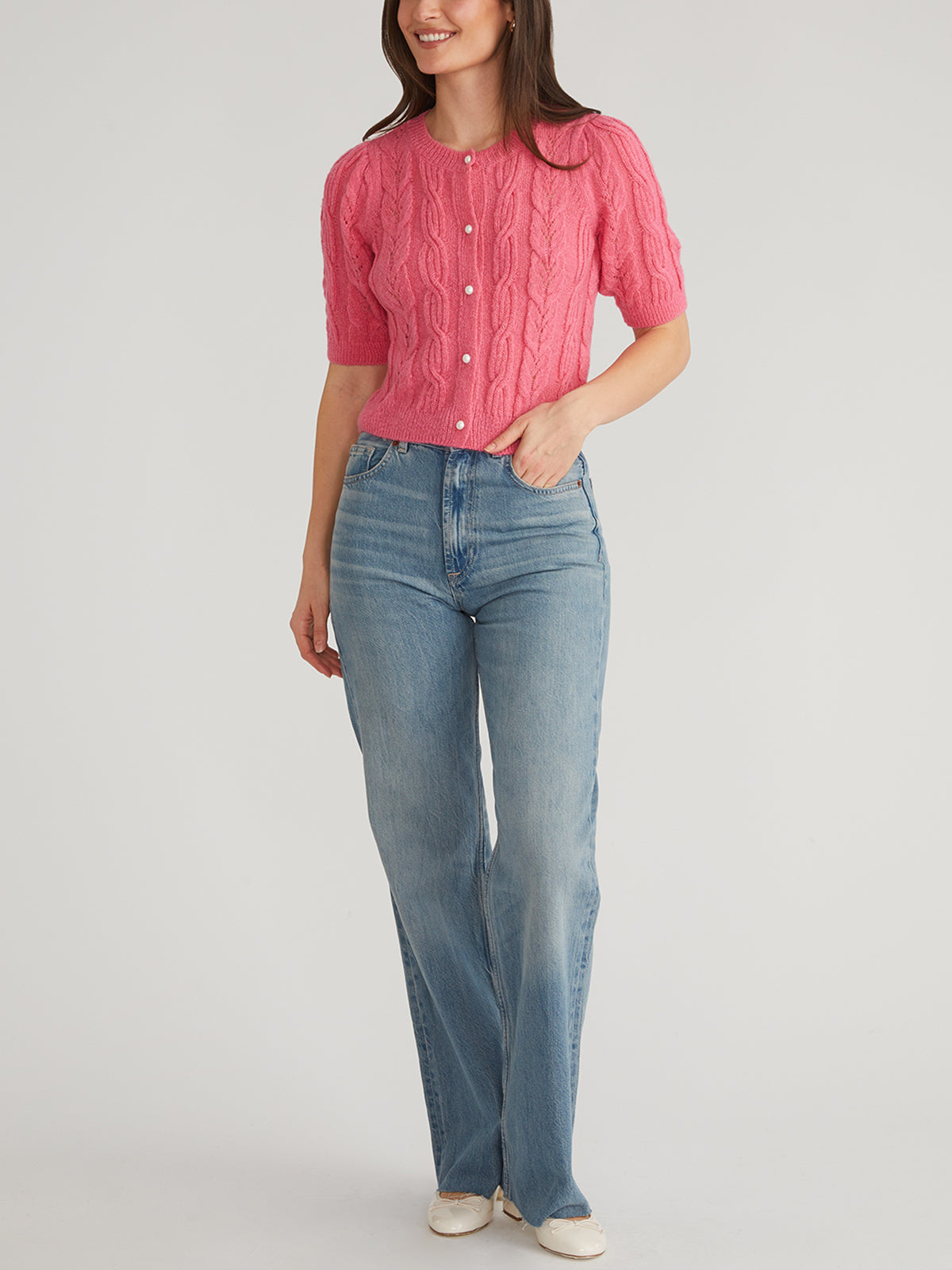 A person stands against a plain background, smiling with their hands by their side, wearing 525 Americas pink cable-knit crewneck cardigan with pearl-like buttons and puff short sleeves, paired with blue high-waisted jeans and white shoes.