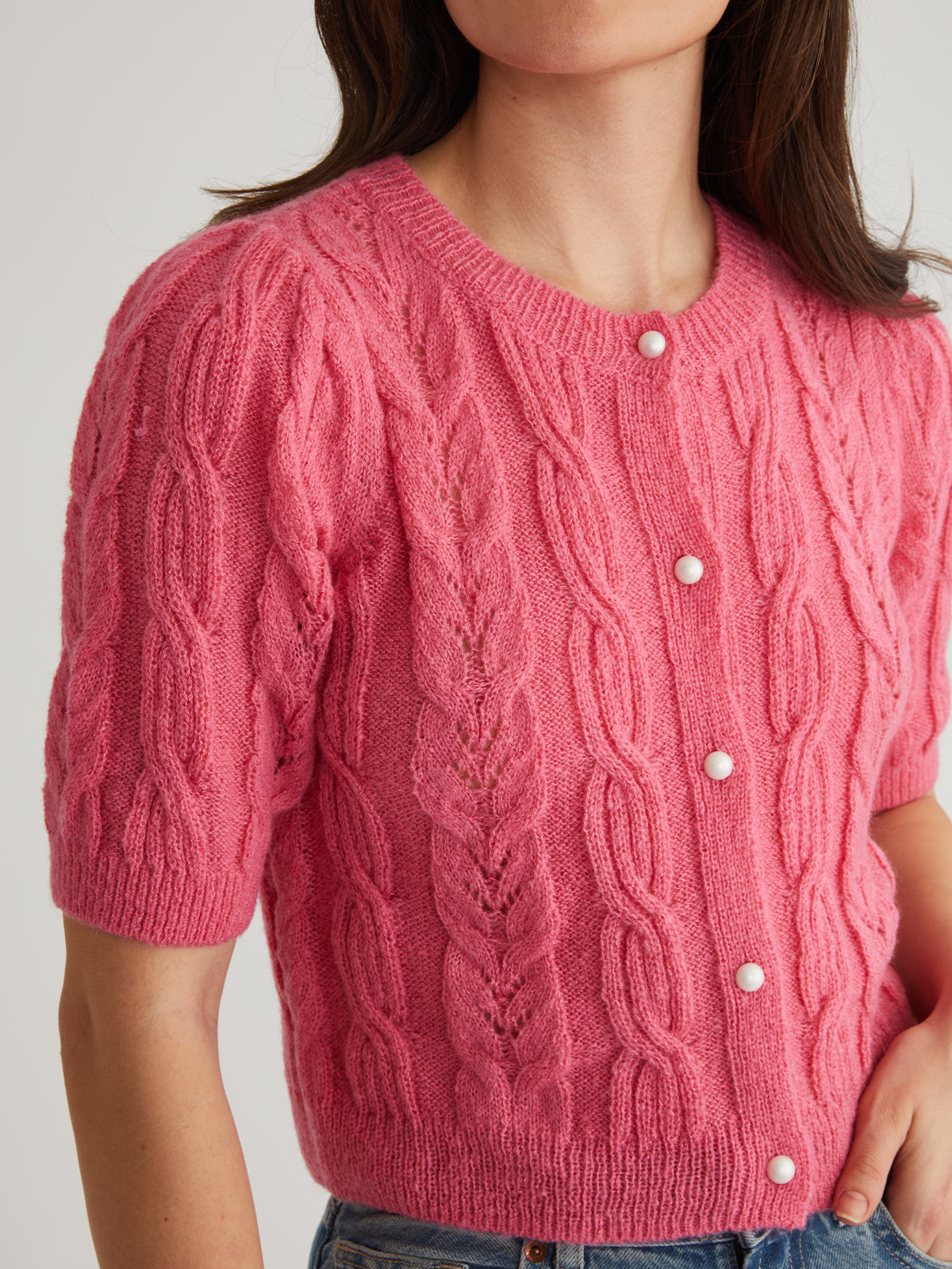 A person poses with one hand on their hip, showcasing a 525 America pink cable-knit cardigan with puff short sleeves, pearl-like buttons, and a charming leaf pattern, stylishly paired with blue jeans.