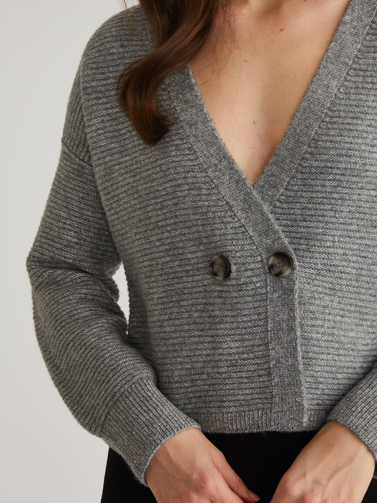 A person is in a gray V-neck 525 America cardigan featuring an ottoman stitch, two dark buttons, long sleeves, and a ribbed texture. They have long, wavy hair with hands near the waist.