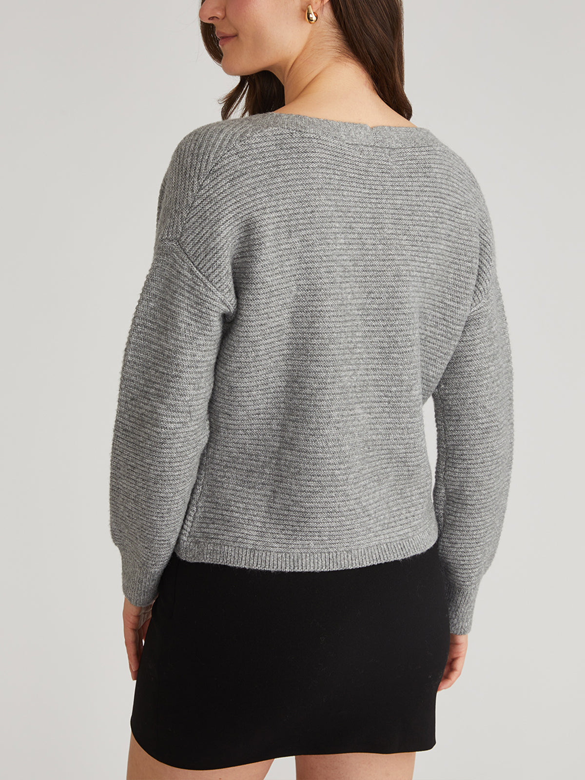 The individual, with long hair and gold hoop earrings, is wearing a gray knitted V-neck cardigan by 525 America paired with a black skirt. They are standing facing away against a plain light gray background.