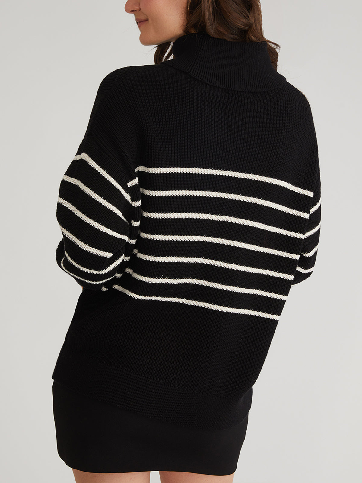 The person is wearing an oversized stripe turtleneck pullover sweater from 525 America, made from 100% cotton. Long brown hair flows down their back as they stand with hands in pockets against a plain light gray background.