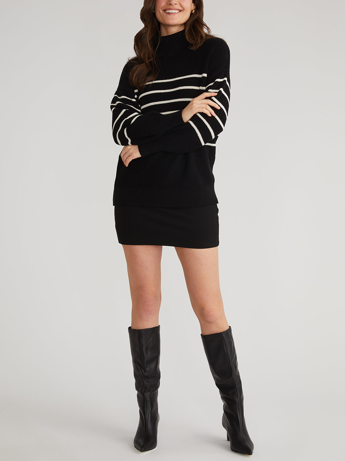 The individual elegantly wears a 525 America oversized stripe turtleneck made of 100% cotton, paired with a black skirt. Their arms are crossed and they smile, finishing the look with knee-high black boots against a light backdrop.