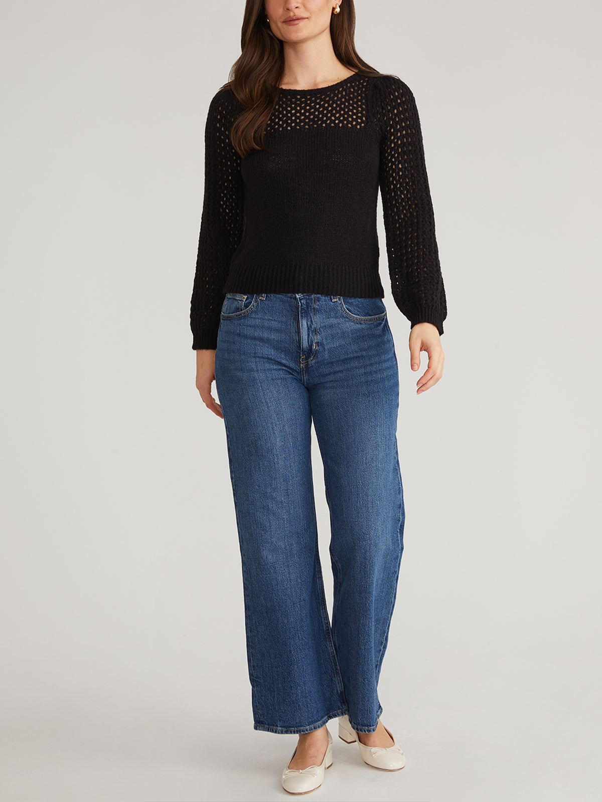 A person with long hair dons a black, puff-sleeved, long-sleeve sweater from 525 America and blue jeans. Against a plain background, their arms relax by their sides. The look is completed with light-colored shoes for modern flair.