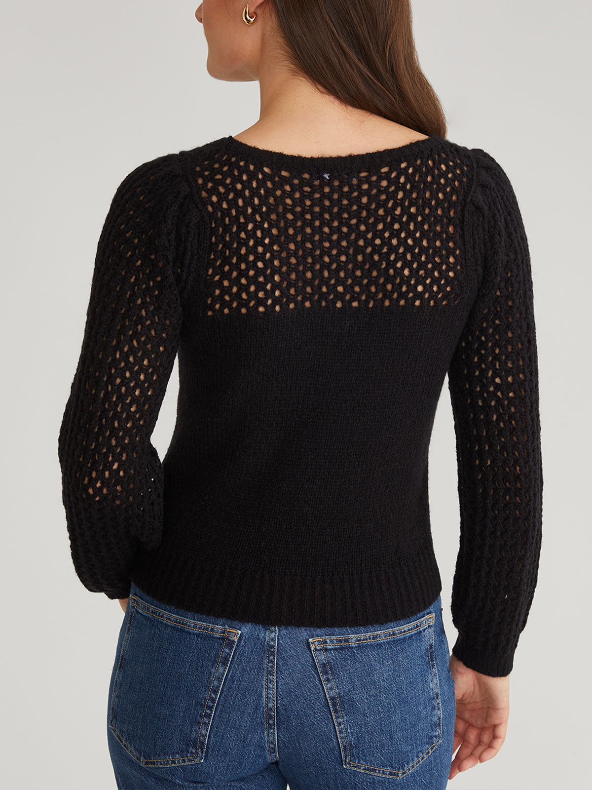A person with long brown hair wears a 525 America black, long-sleeved crochet sweater with modern mesh detailing on the upper back, paired with blue jeans. The image captures a back view of the outfit against a light gray background.