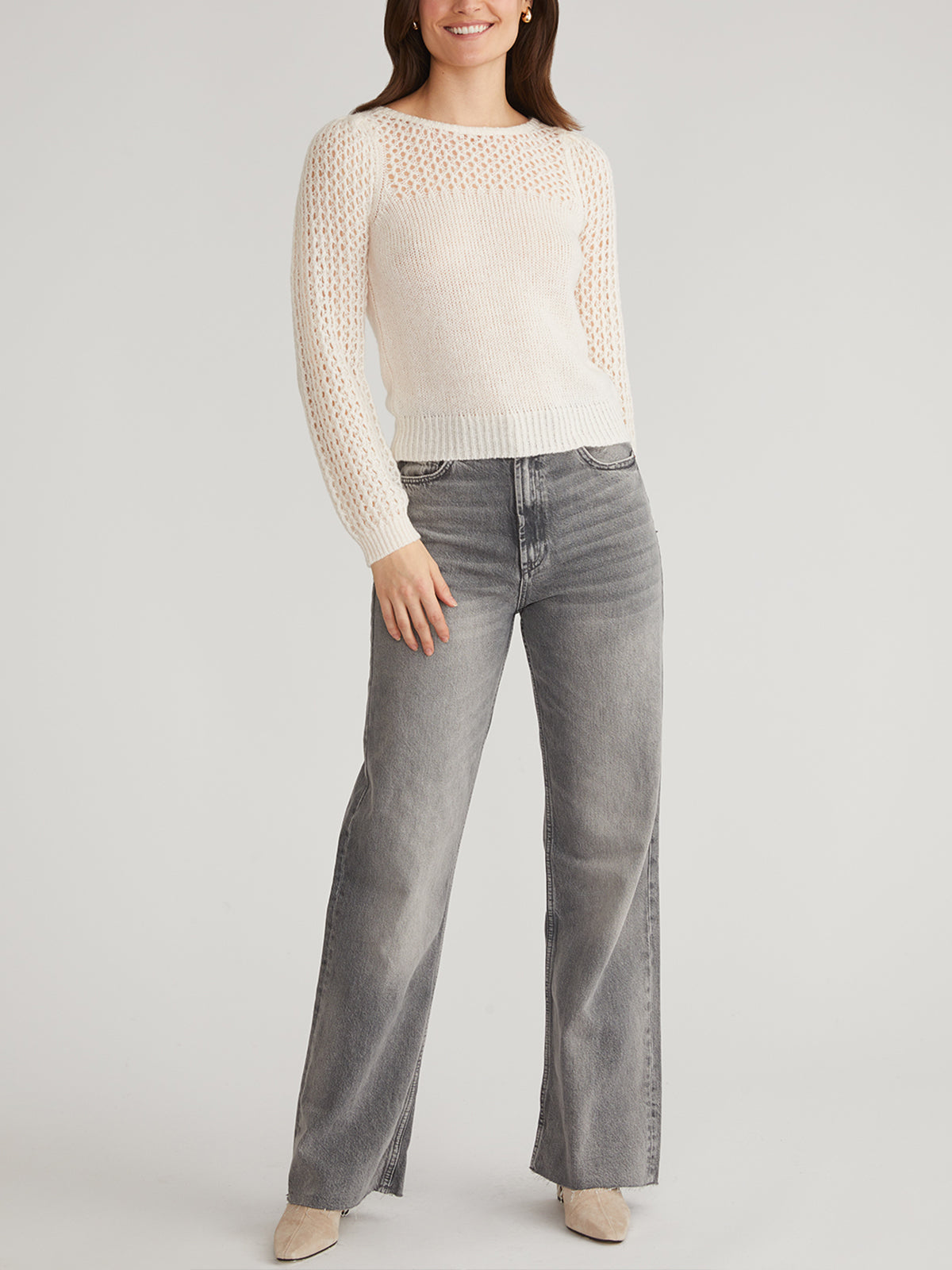 A person wearing a white 525 America knit sweater with a lattice pattern and puff sleeves pairs it with gray wide-leg jeans and beige shoes, standing against a light background with a slight smile and relaxed arms.