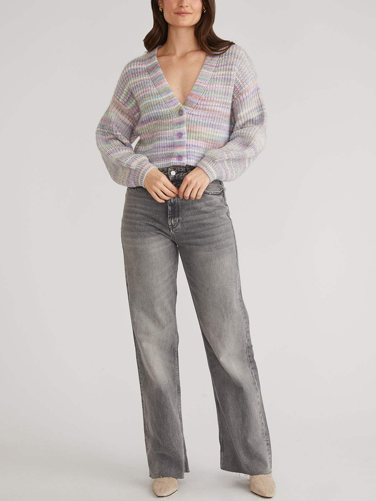 Against a plain backdrop, a person models a light-striped, V-neck Libby Cardigan with balloon sleeves from 525 America, paired with gray jeans and beige shoes.