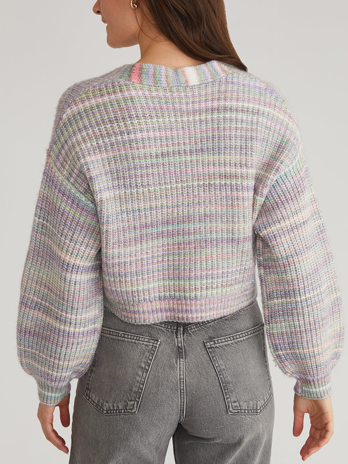 A person with long hair models a pastel-colored plaid-knit sweater from 525 America, crafted from spacedye fabric with a round neckline and balloon sleeves. Paired with high-waisted gray jeans, they are turned away, highlighting the sweaters back design.