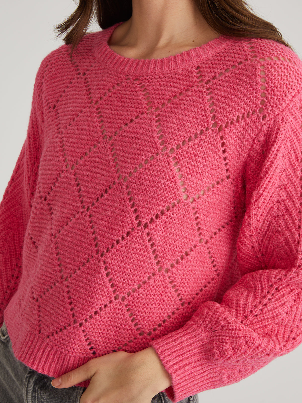A person wears a bright pink 525 America pointelle pullover with a diamond pattern, featuring balloon sleeves and a textured look. Part of their face is visible as they casually tuck one hand into their jeans pocket.