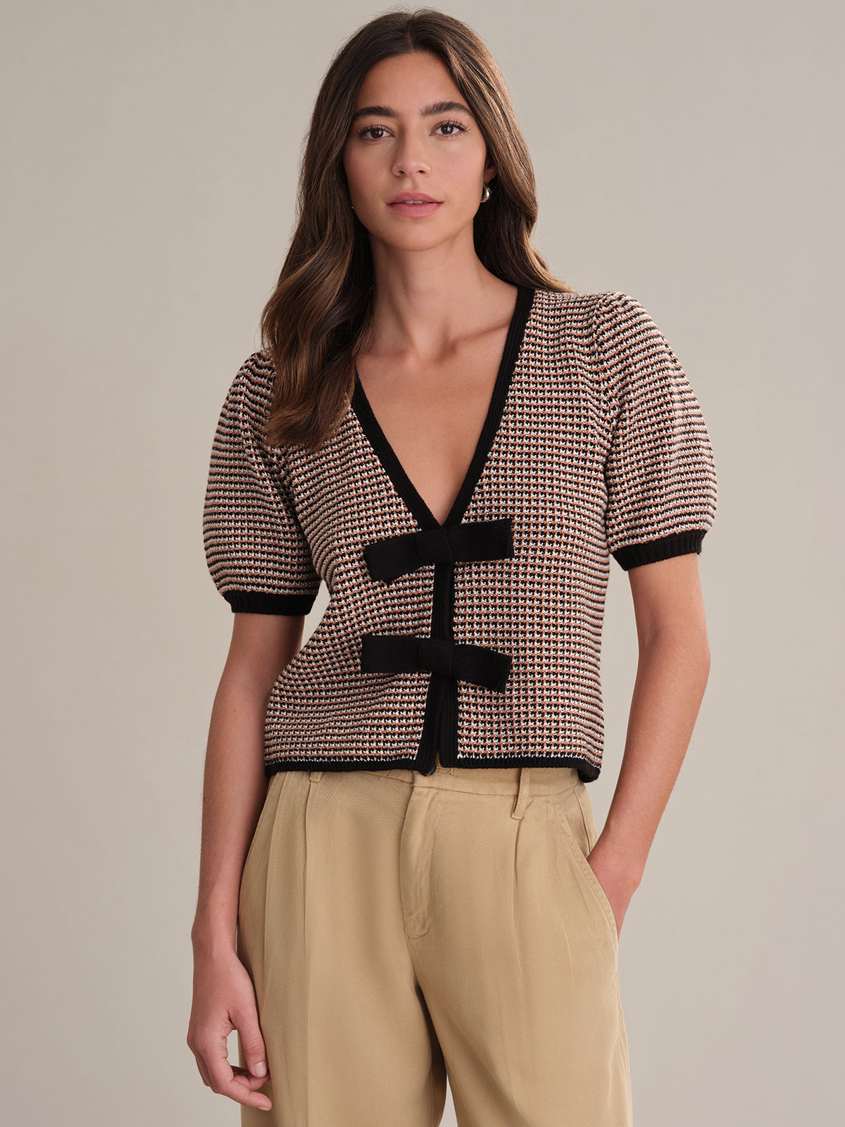 A woman with long brown hair models a 525 America 100% cotton feminine cardigan with a houndstooth pattern and two black bow ties, paired with high-waisted tan trousers, standing against a plain beige background.