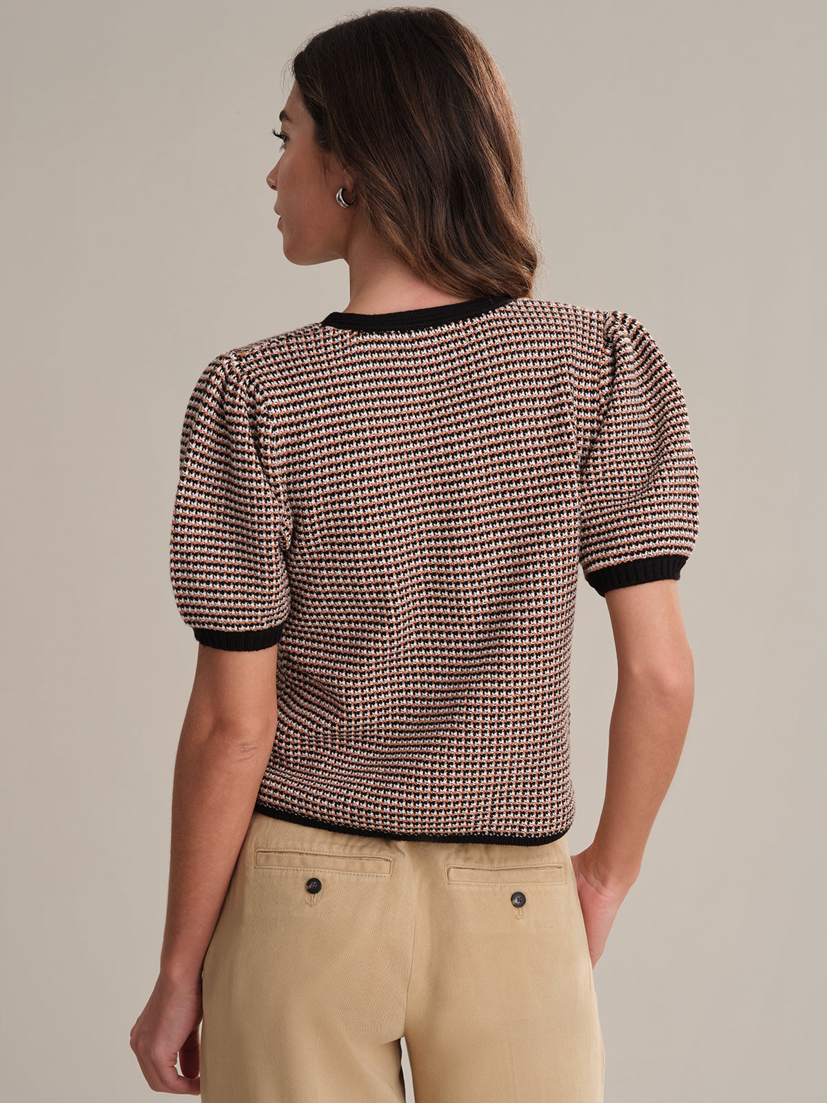 A woman with shoulder-length brown hair faces away, wearing a 525 America short-sleeved textured knit top with black trim, beige pants, and her left hand in her pocket. The 100% cotton top stands out against a plain background.