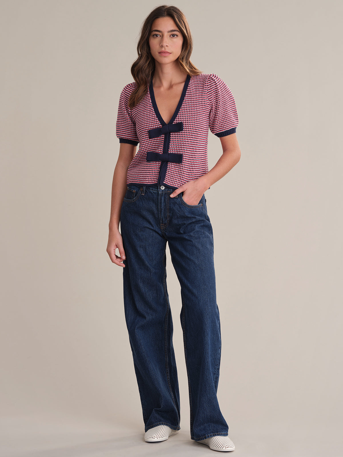 A person stands against a plain background wearing a 525 America short-sleeve cardigan with red and navy checks, black trim, and toggle buttons. They pair it with loose blue jeans and white shoes, with one hand in their pocket.