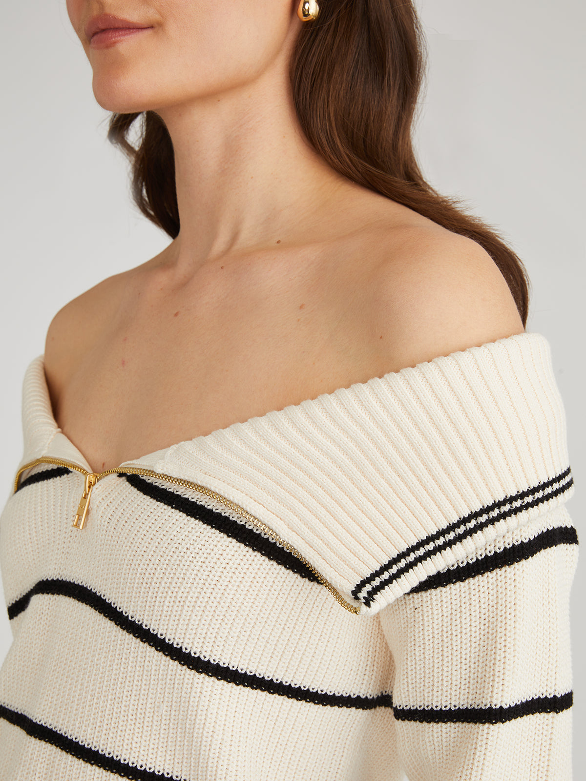 Wearing a 525 America off-the-shoulder striped pullover with an adjustable zippered neckline, a person with brown hair is highlighted by a small gold earring against a softly lit, plain background.