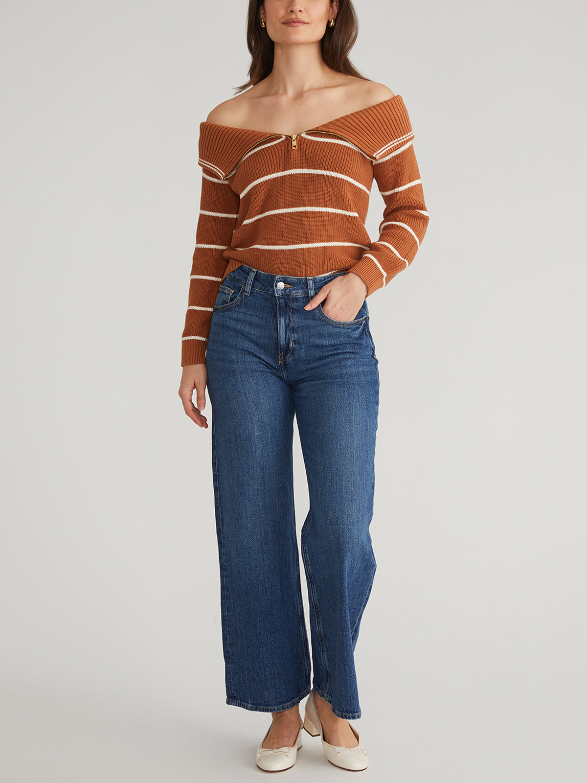 A person models a brown, striped off-the-shoulder 525 America pullover sweater paired with blue wide-leg jeans. Their hand rests in their pocket, and they wear white shoes. The 100% cotton outfit is set against a light-colored background.