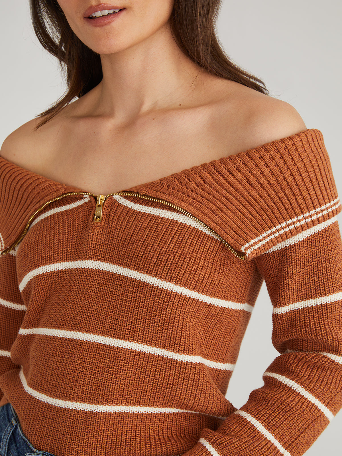 A person is wearing a brown striped, off-the-shoulder pullover from 525 America. Made from 100% cotton, it features an adjustable zippered neckline and pairs effortlessly with blue jeans against a neutral background.