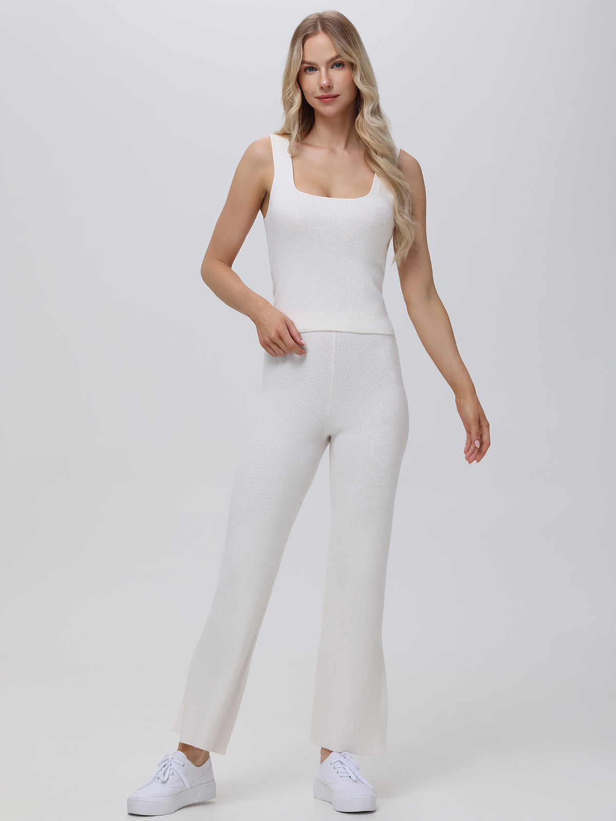 A person with long blonde hair poses confidently against a light gray background, dressed in a versatile sleeveless white top and form-fitting matching pants from 525 America. They complete the look with white sneakers and rest their right hand on their hip while gazing forward.