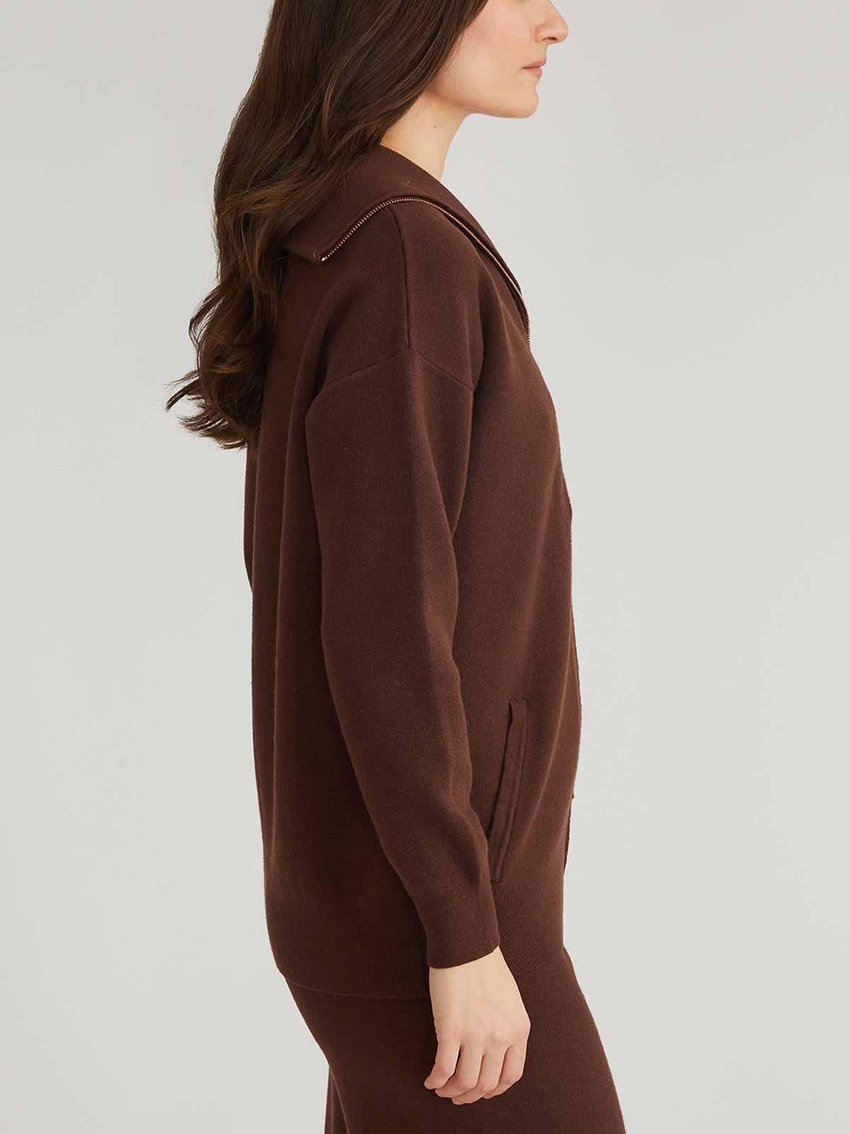 A person with long brown hair stands in profile against a plain light background, wearing a 525 America luxe yarn knit zip front cardigan sweater with an oversized collar, along with matching pants. Their hands are relaxed at their sides.