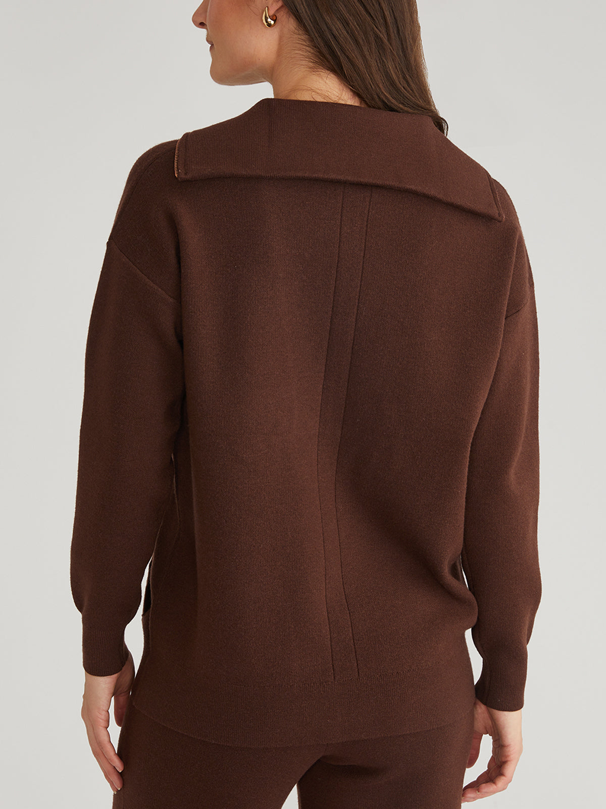 A person with long hair is facing away, wearing a 525 America brown sweater and pants set. The oversized sweater, made from luxe yarn, has a wide collar and vertical seam on the back. The background is plain and light-colored.